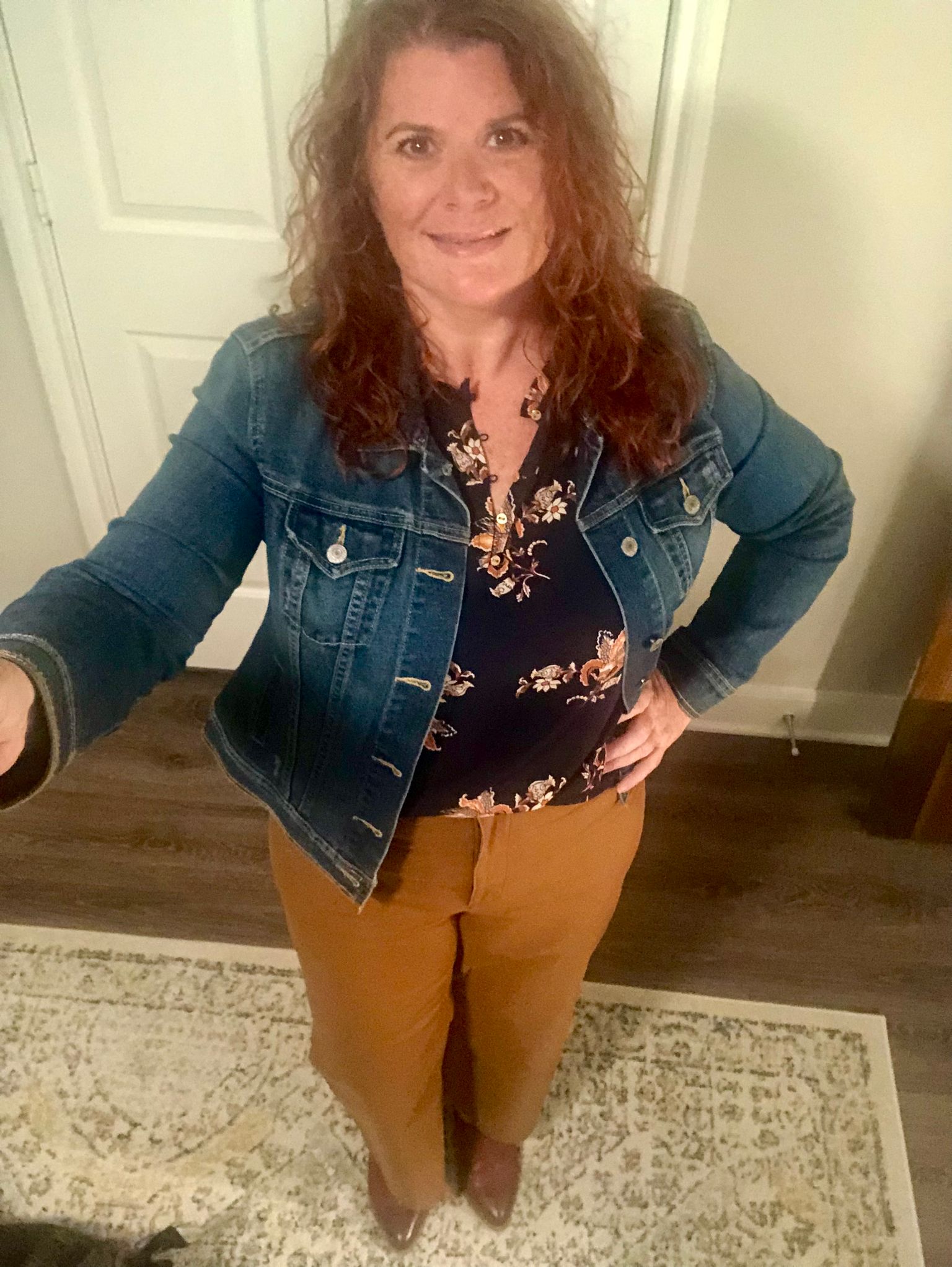 Smiling woman taking a picture of herself wearing a denim jacket, black floral blouse and mustard pants
