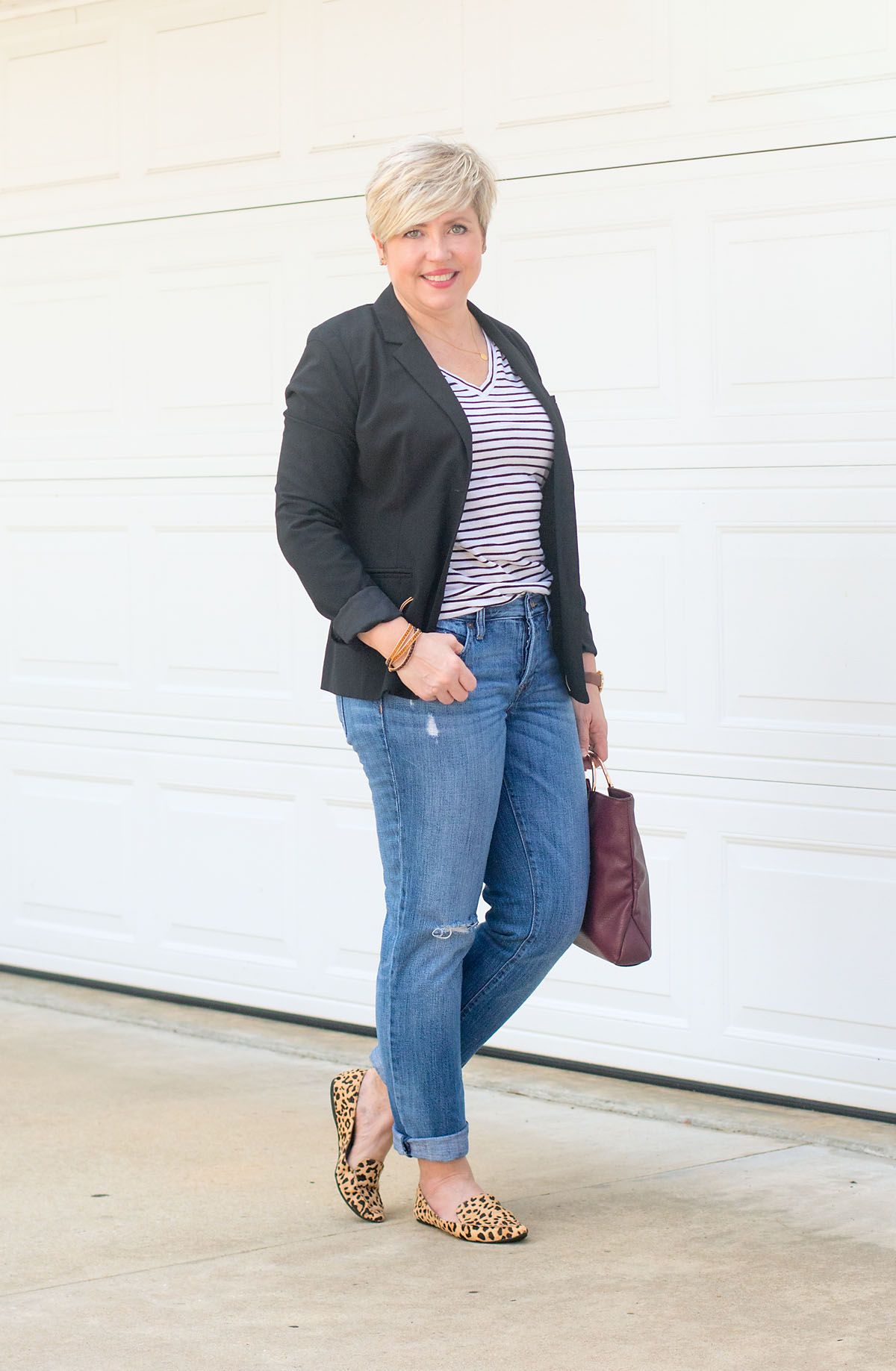 Woman posing in front of a garage door wearing a black blazer, black and white stripe shirt, jeans, and leopard flats while holding brown purse
