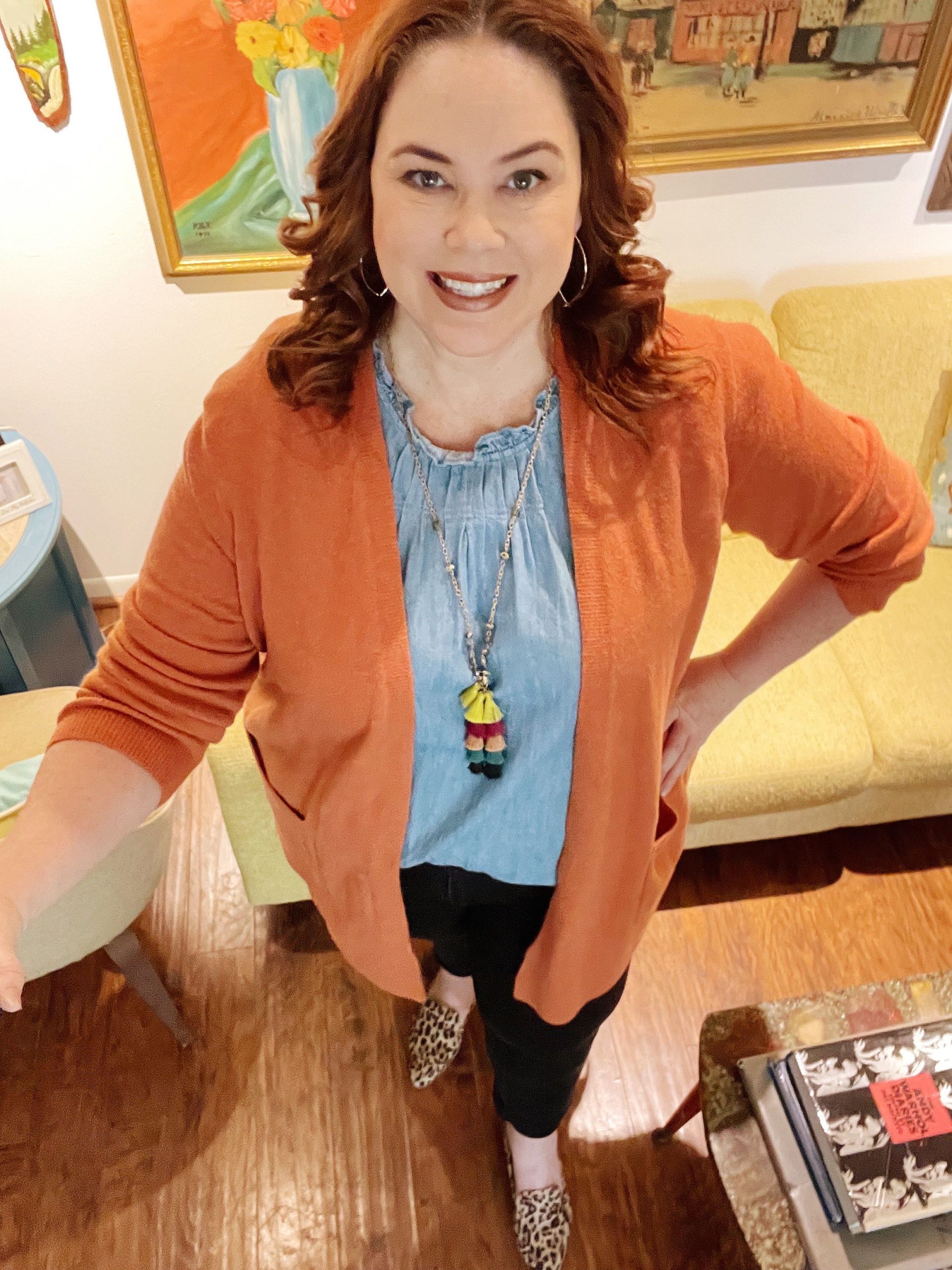 Woman posing in her living room, wearing a orange sweater, chambray top, statement necklace, black pants, and animal print shoes