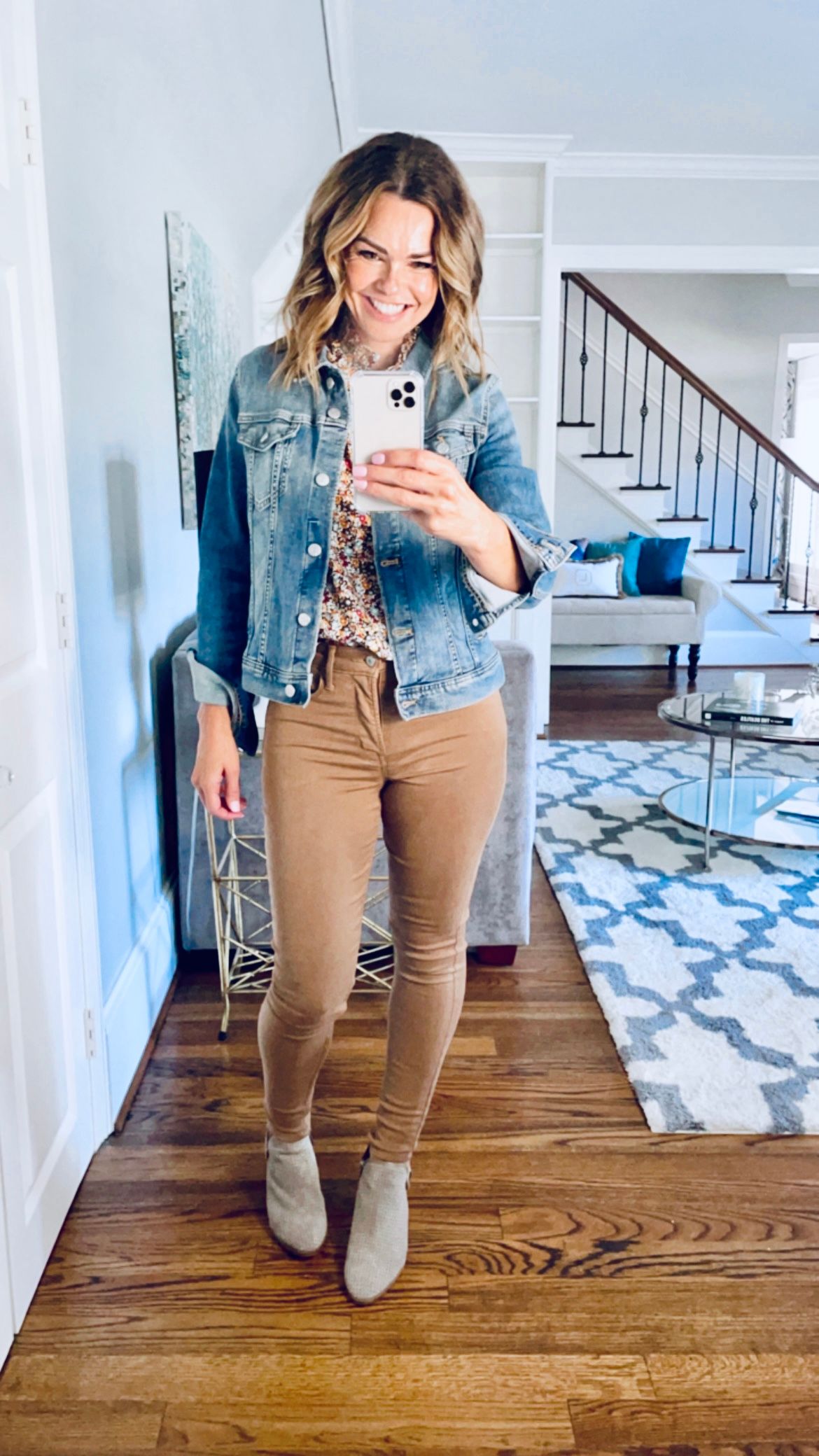 Smiling woman taking a full body selfie of her outfit - denim jacket, floral shirt, brown skinny corduroys and grey booties