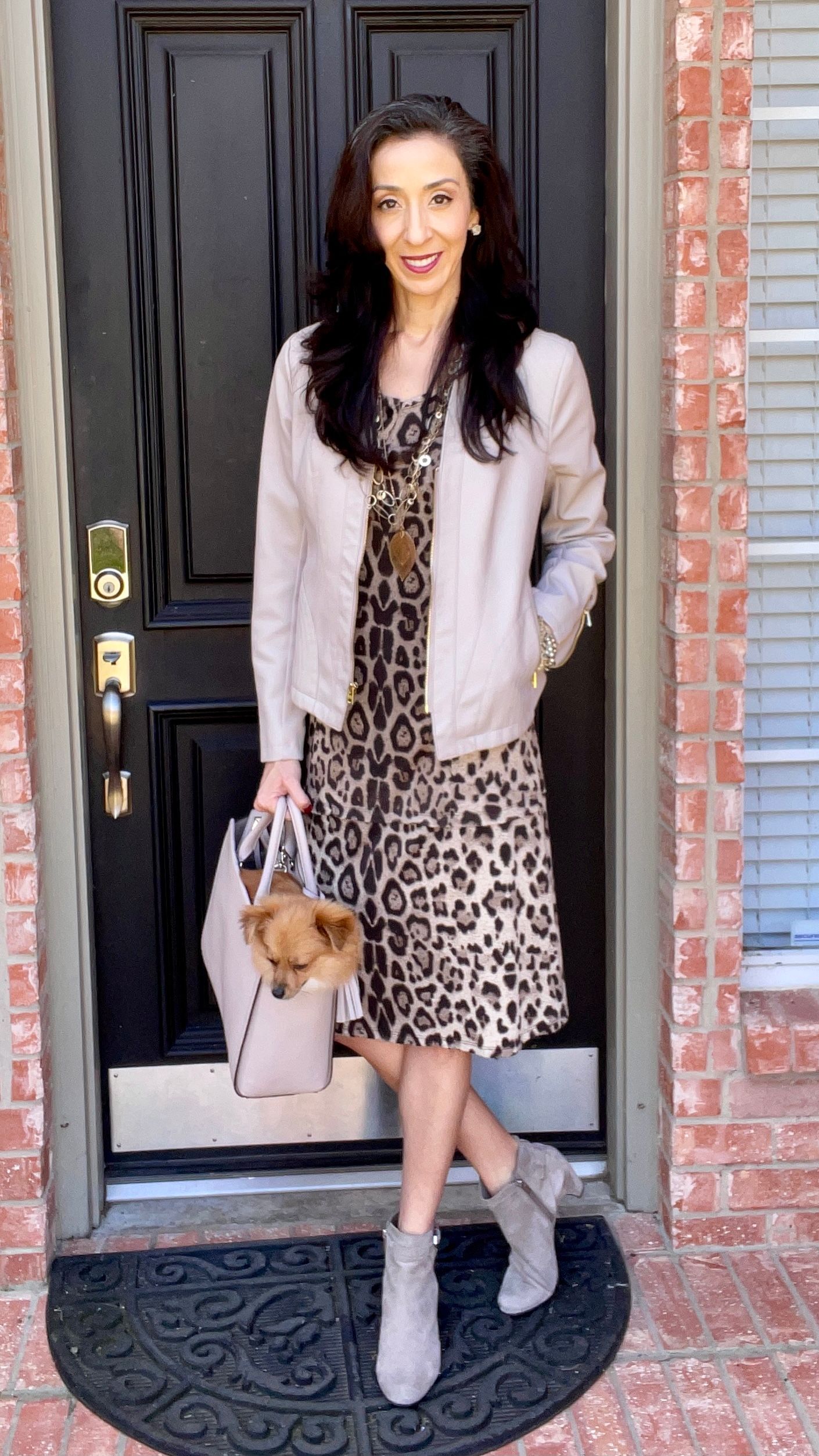 Smiling woman with brunette hair posing in front of her home, holding a bag with a small dog inside, wearing an animal print knee-length dress, taupe leather jacket, and gray booties