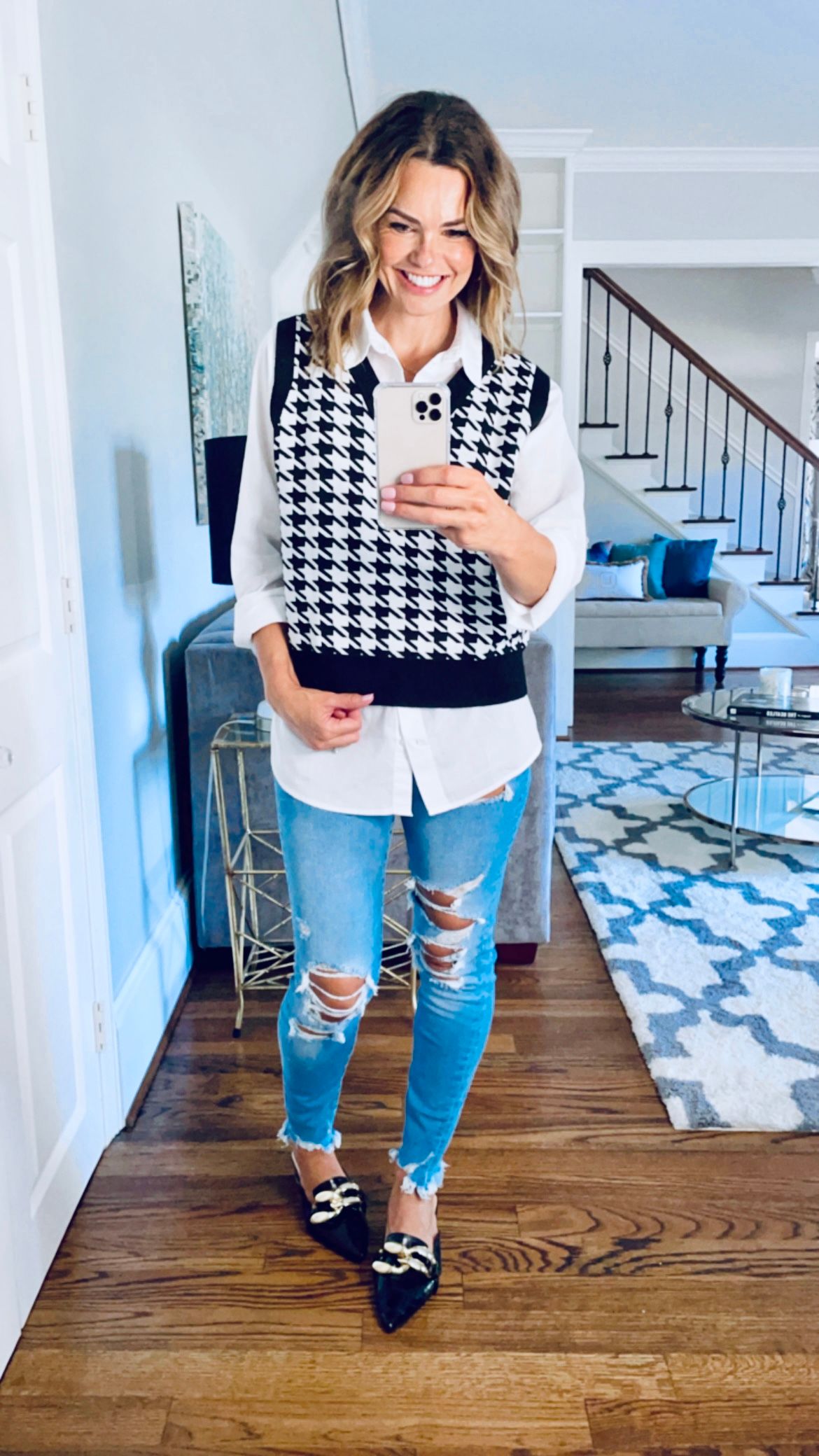 Beaming woman taking a full body selfie in her living room, wearing a white button down shirt with houndstooth sweater over it, denim ripped jeans, and black pointed toe flats