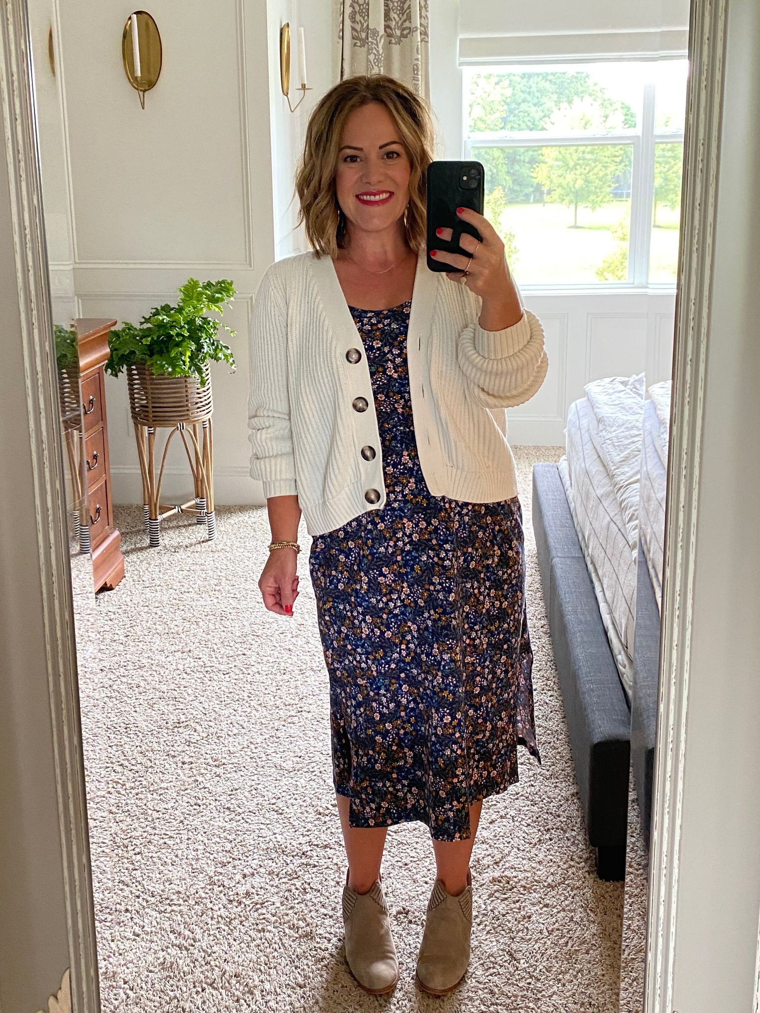 Happy woman taking a full body selfie in her bedroom, wearing a floral midi dress, white sweater, an grey booties