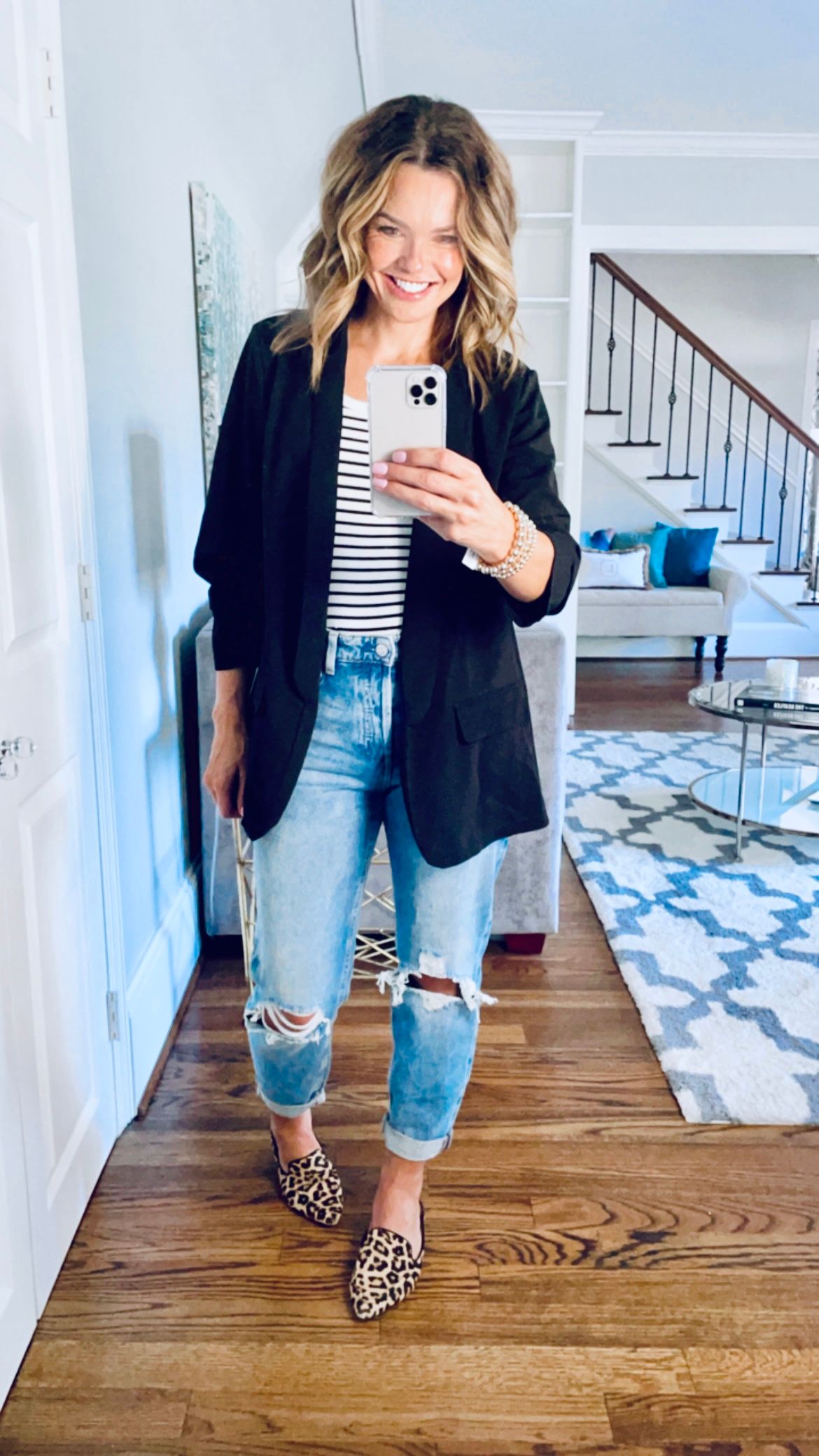Smiling woman taking a full body selfie while wearing a black blazer, black and white stripe shirt, denim ripped jeans, and animal flats