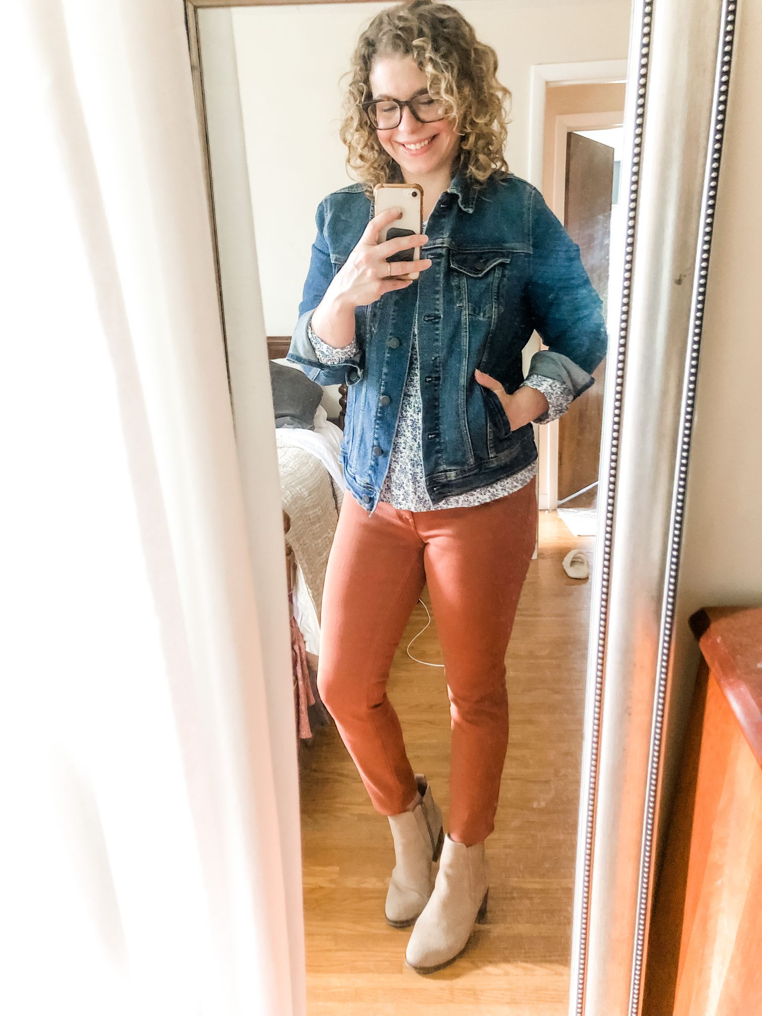 Smiling woman with blonde curly hair and glasses taking a full body selfie of her outfit - denim jacket, floral long sleeve shirt, orange pants and khaki boots