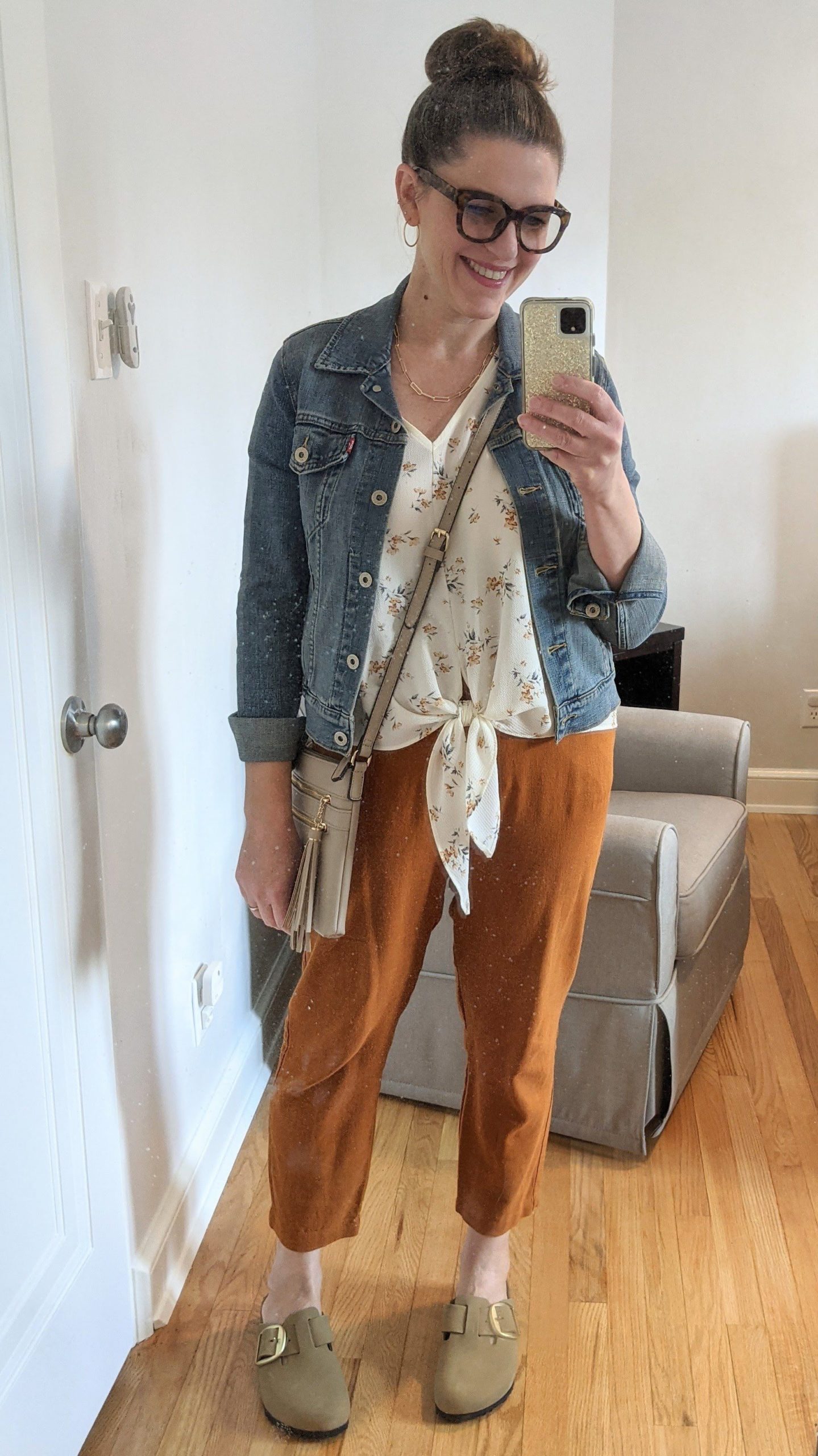 Smiling woman with brunette hair in a bun and glasses taking a full body selfie of her outfit - denim jacket, floral tie blouse, orange pants and flats