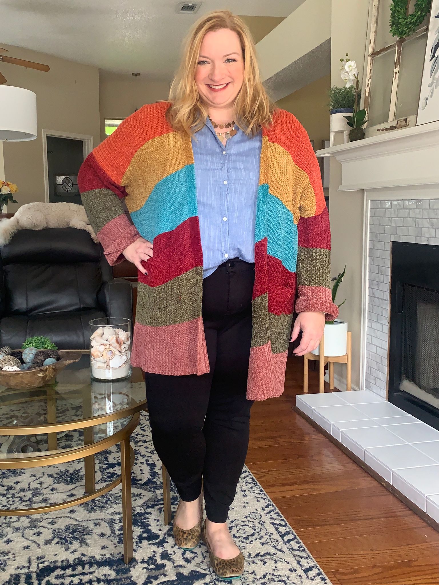 Plus size woman posing in her living room, wearing a chambray button down shirt, multi colored/striped sweater, black pants, and animal print flats.