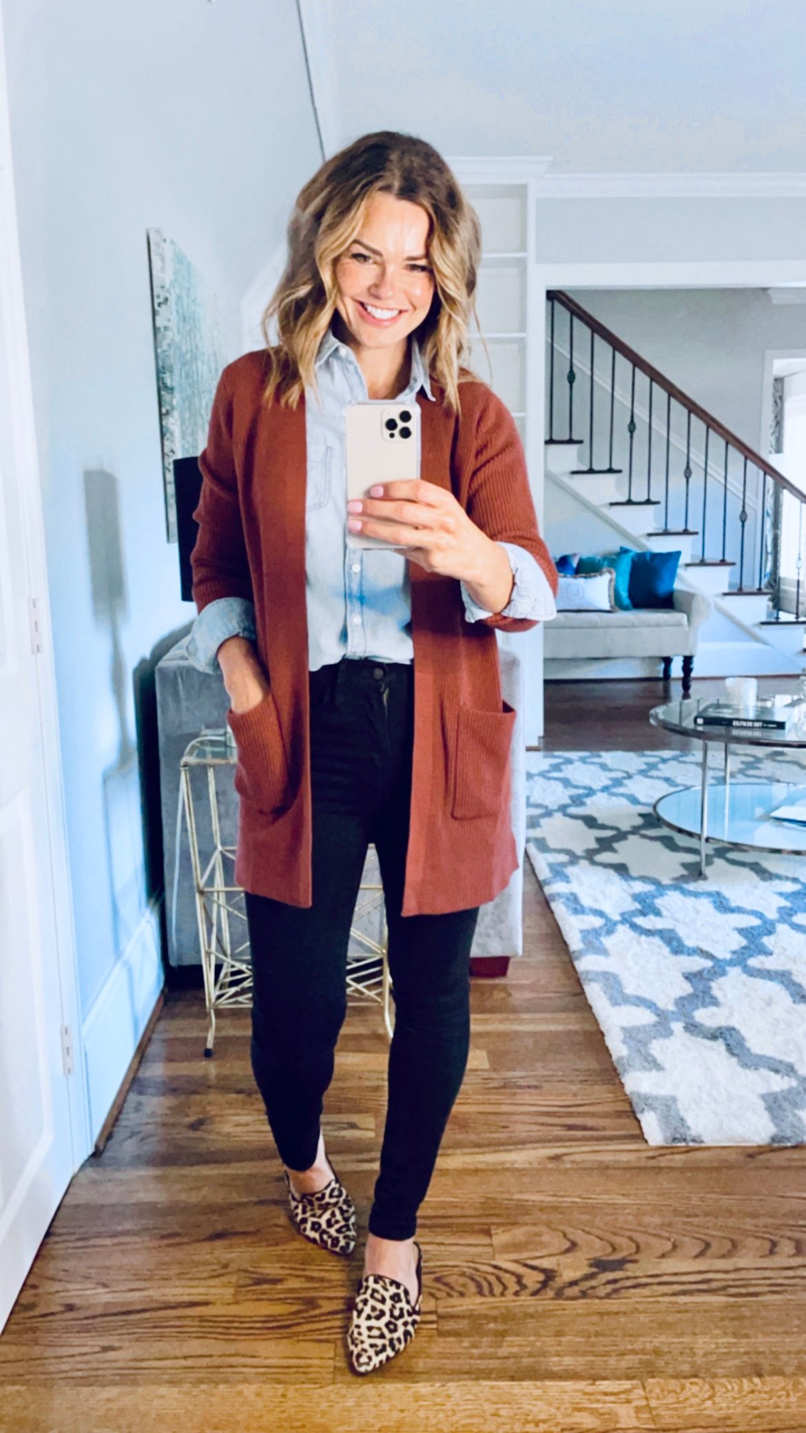Happy woman taking a full body selfie in her living room, wearing a chambray button down long sleeve shirt tucked into black skinny jeans, with a warm brown sweater and animal print shoes