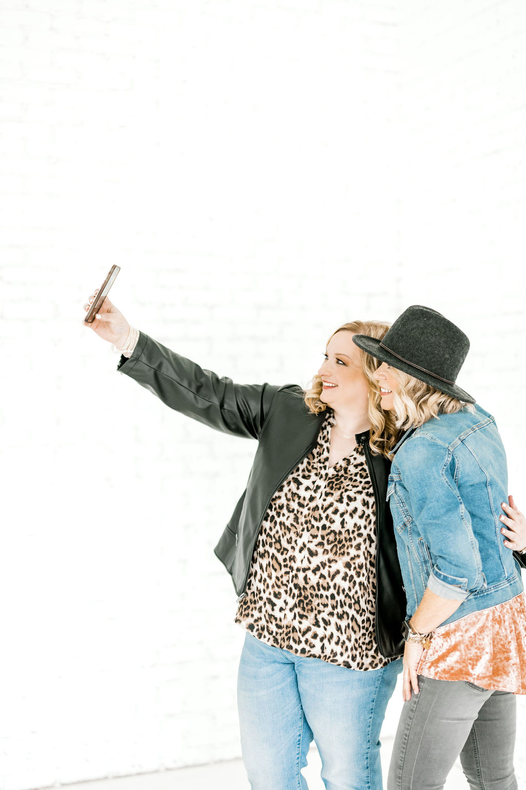 Two women taking a selfie together - one woman is wearing an animal print top, black leather jacket and jeans and the other woman is wearing a charcoal hat, denim jacket, orange top and grey jeans